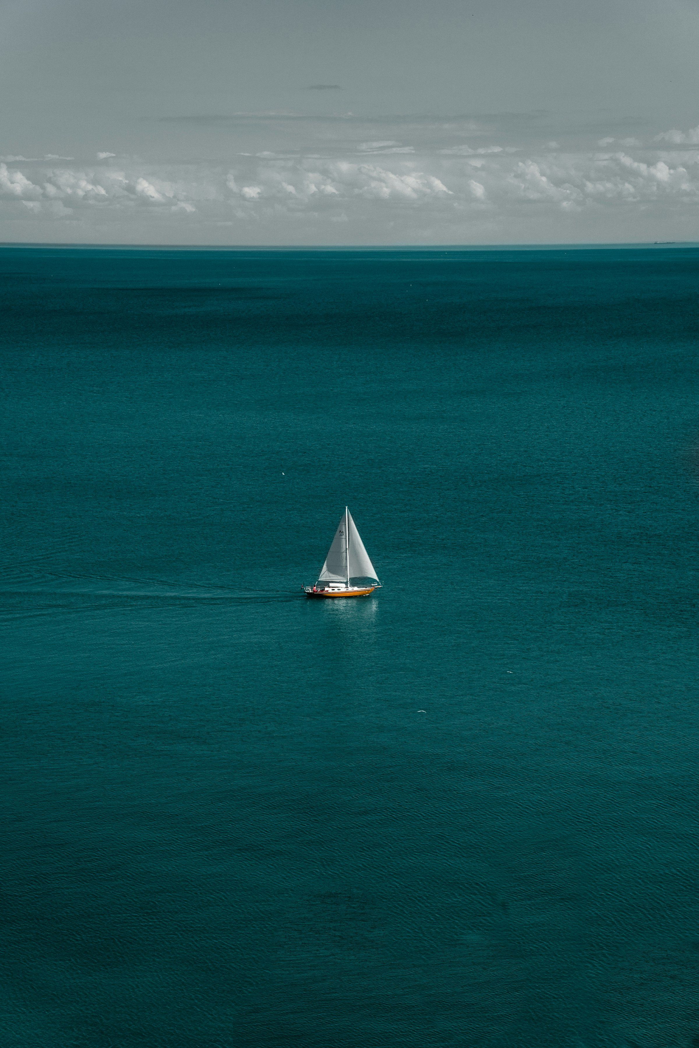 A lone sailboat crossing a wide, deep-teal sea beneath a cloudy sky.